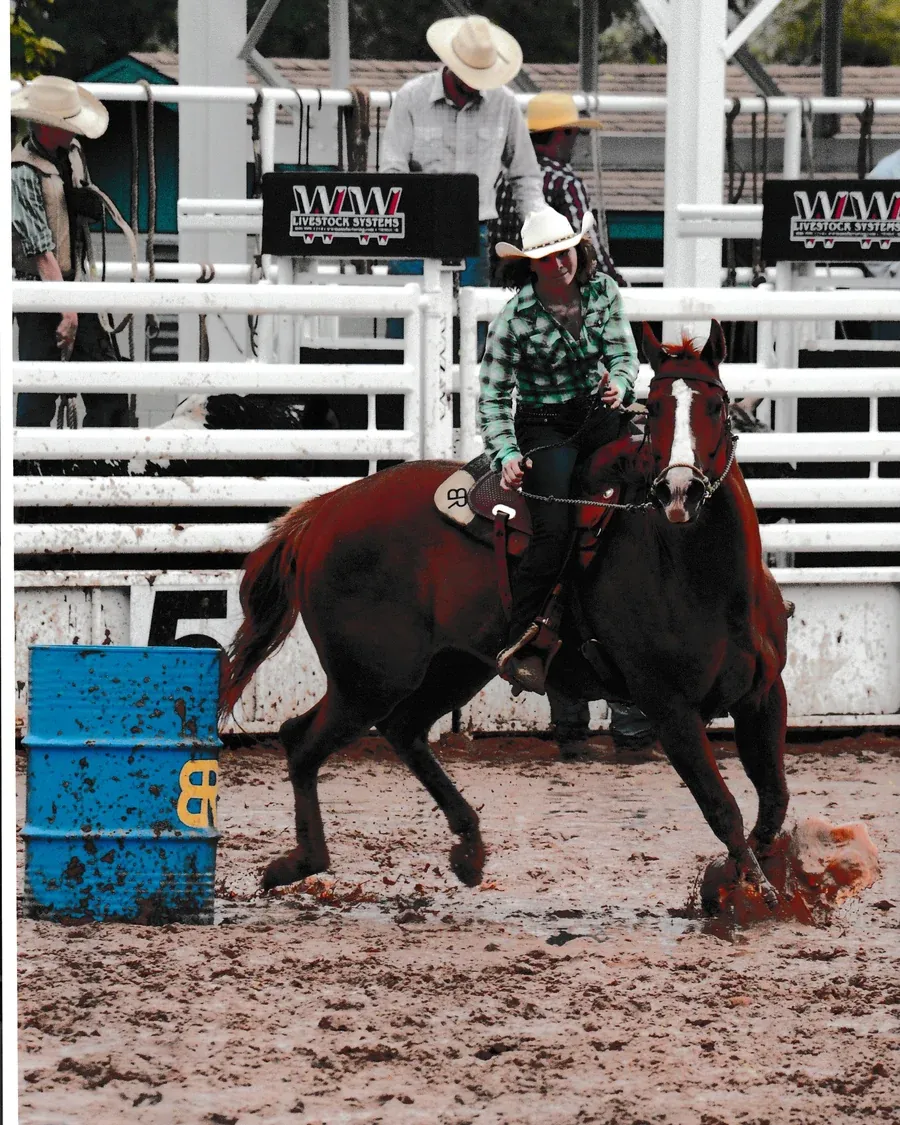 Loren rounding a barrel on a dark bay horse in a wet arena at the Boys Ranch Rodeo, BR branded on the saddle pad and the barrel